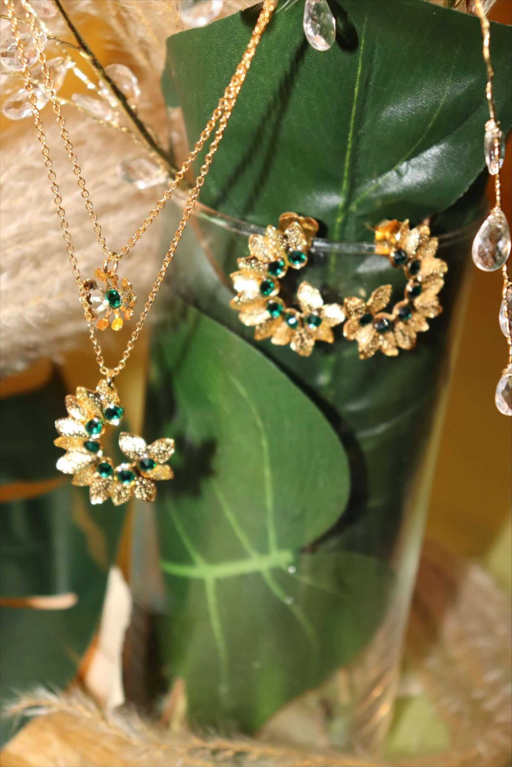 Gold floral necklace and earrings with green gemstones displayed on a green leaf