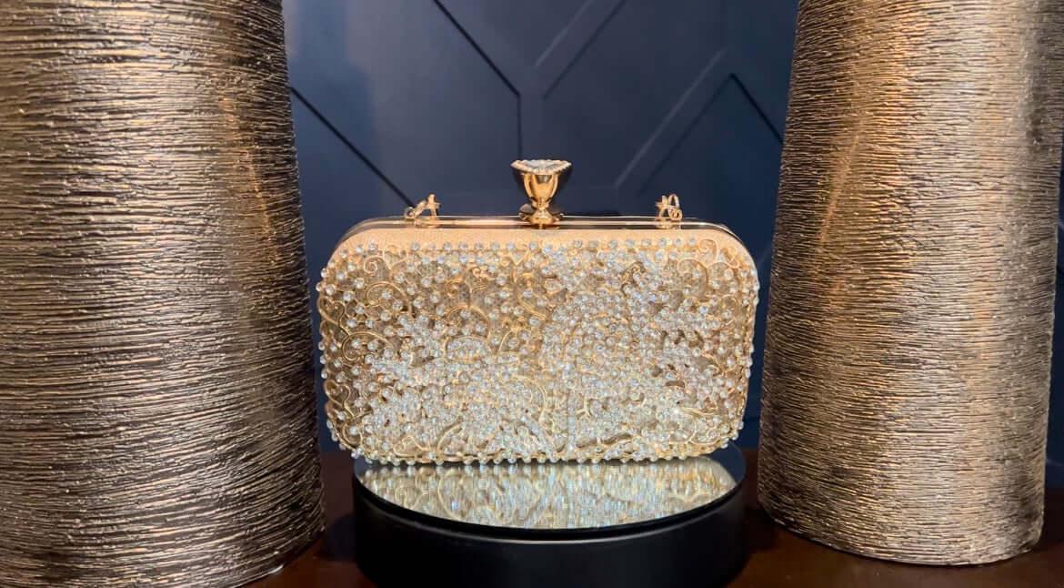Gold crystal-embellished clutch purse displayed on a mirrored stand with textured gold vases