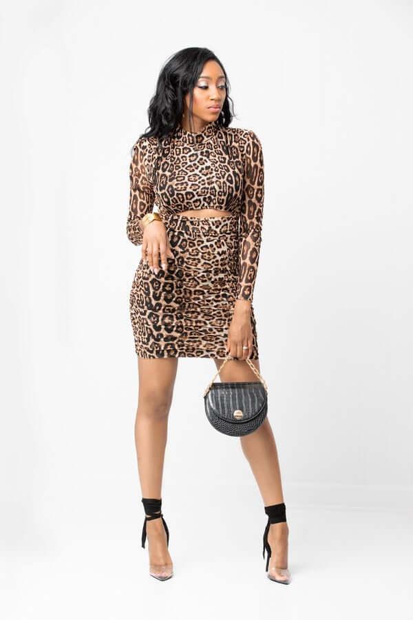 Woman in leopard print bodycon dress with black heels and round black handbag on white background