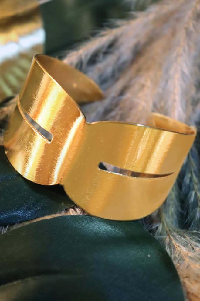 Brushed gold metal cuff bracelet with cutout design on green and beige fabric background